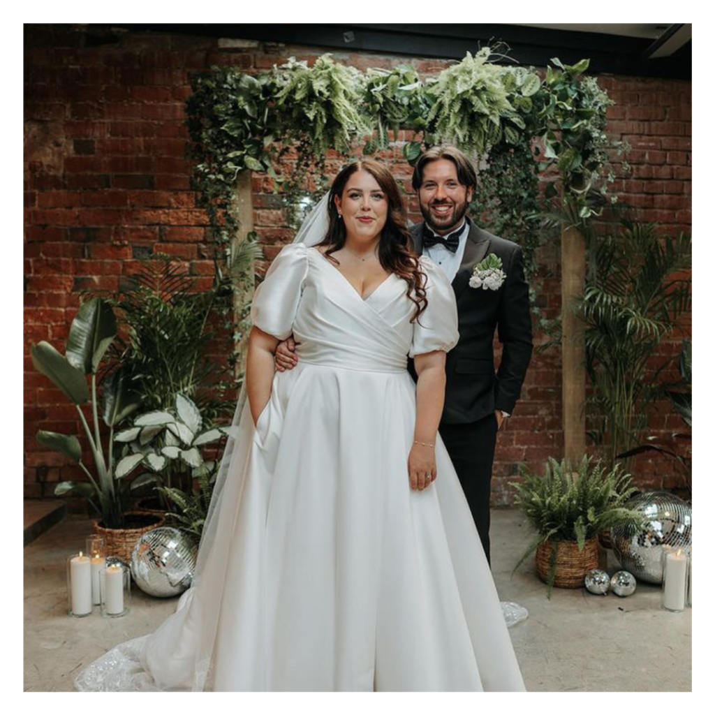 Bride and groom standing together beneath a greenery arch with disco balls and brick backdrop at The Shack Revolution