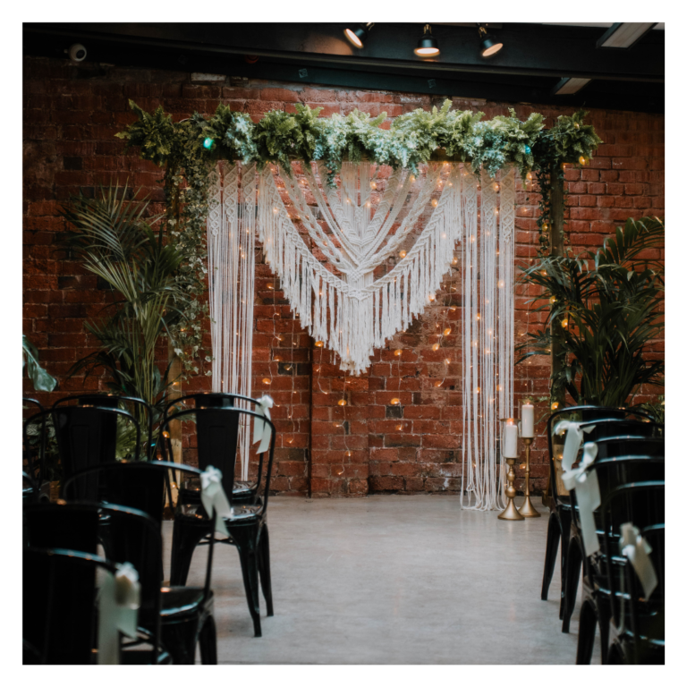 Living foliage ceremony backdrop with macramé draping against an exposed brick wall at The Shack Revolution
