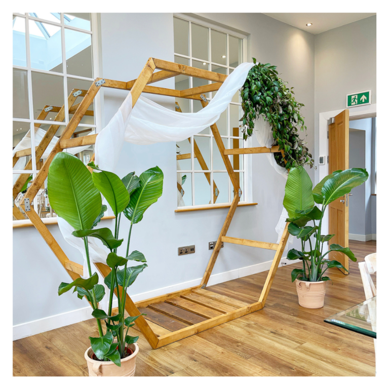 Side view of a hexagon wooden wedding arch with white fabric draping and lush greener