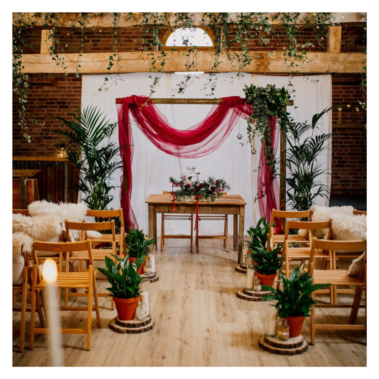Living plant wedding ceremony backdrop with burgundy draping and potted greenery at The Haybarn