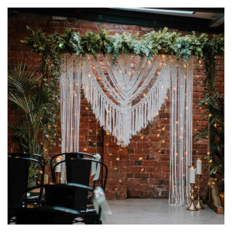 Boho macramé wedding backdrop with fairy lights and greenery against a brick wall at The Shack Revolution wedding venue in Hereford.