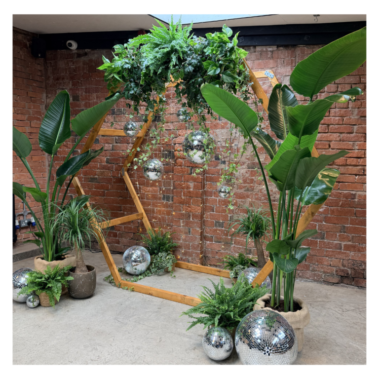Living plant wedding backdrop featuring a wooden hexagon arch, tropical greenery, disco balls and trailing foliage for alternative wedding styling.
