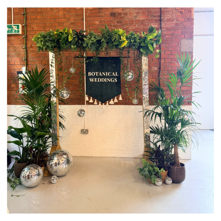 Botanical wedding photo backdrop featuring disco balls, tropical plants, hanging greenery and modern wedding styling for guest photographs. Disco balls at the feet of it and disco ball tape on the backdrop.