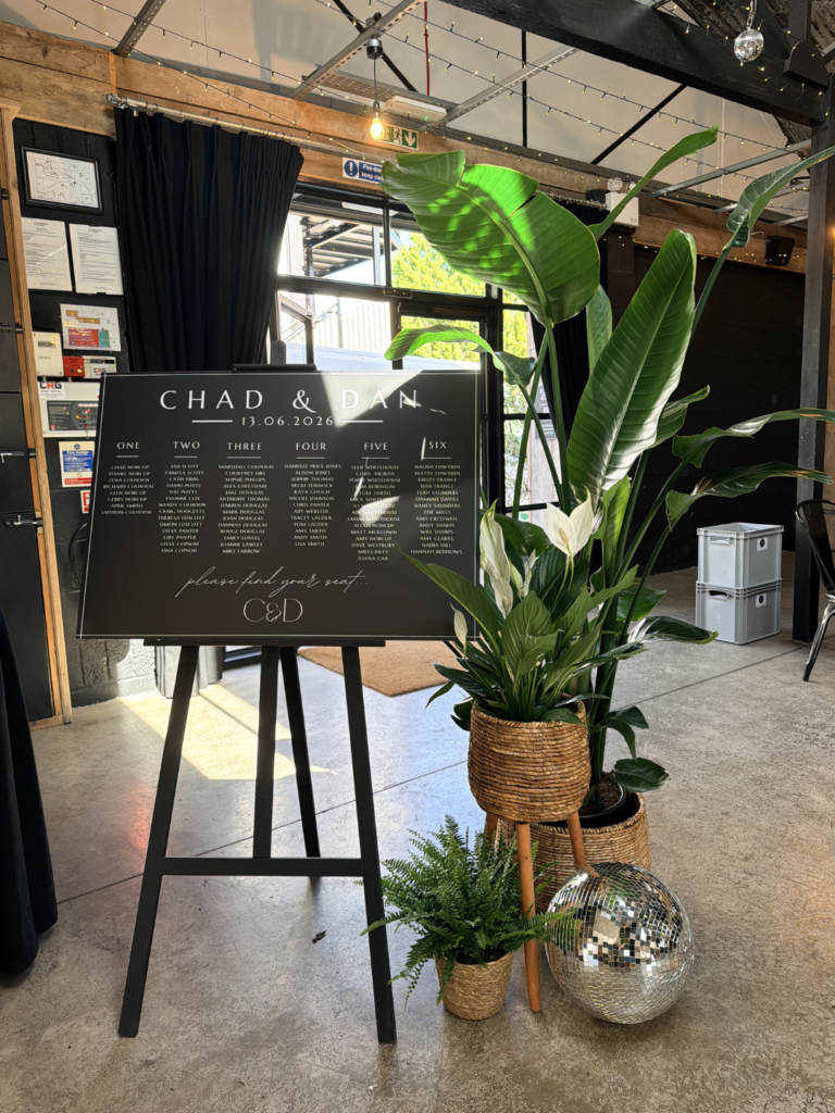 An arrangement of plants next to a seating plan at a wedding. There is a monochrome theme.