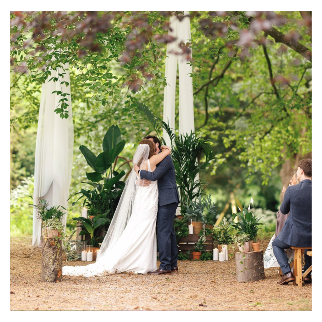 Just married - a couple kiss infront of a living ceremony backdrop made from ferns, creates, palms, candles and long white drapes.