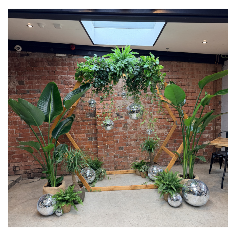 Living plant wedding backdrop featuring a wooden hexagon arch, tropical greenery, disco balls and trailing foliage for alternative wedding styling.