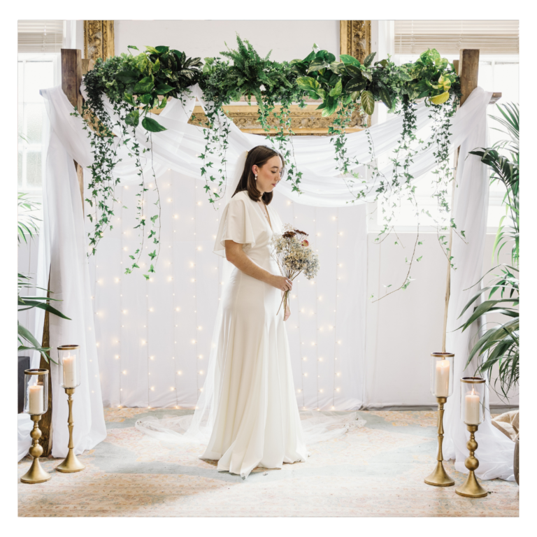 Botanical wedding ceremony backdrop featuring tropical plants, draped fabric, fairy lights and trailing ivy for sustainable wedding décor. Featuring a bride.