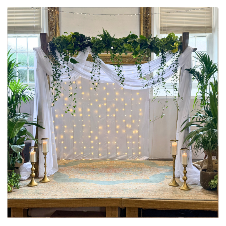 Wedding ceremony backdrop with flowing white drapes, fairy lights and cascading living plants, creating a romantic botanical wedding setting.