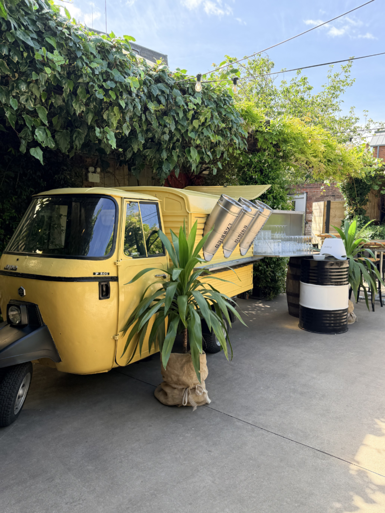 A Yellow Tuk Tuk that dispenses drinks framed by cordylines.