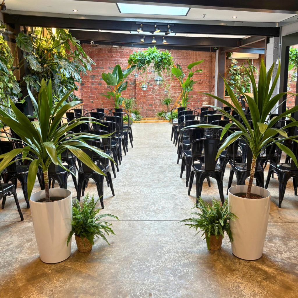 Indoor wedding ceremony with tropical plants framing the aisle and a living foliage backdrop creating a natural botanical wedding setting.