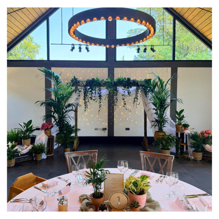 Botanical wedding backdrop with living palms, trailing foliage, soft white draping and fairy lights behind a reception table.