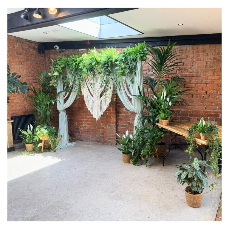 Simple wooden wedding backdrop covered in trailing living foliage and surrounded by palms, peace lilies and smaller potted plants.